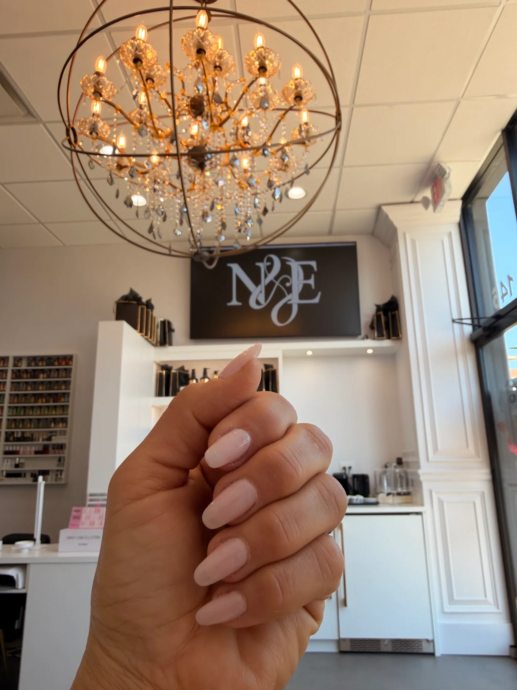 Milky almond manicure held up beneath the crystal orb chandelier and the N&E sign inside Ridgewood Nail Club.